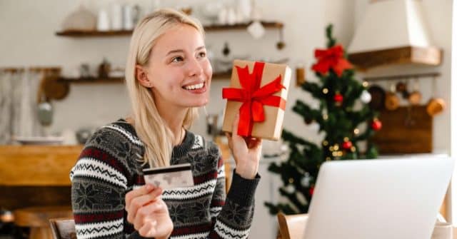 Blonde woman holding credit card and christmas gift with computer and christmas tree in background